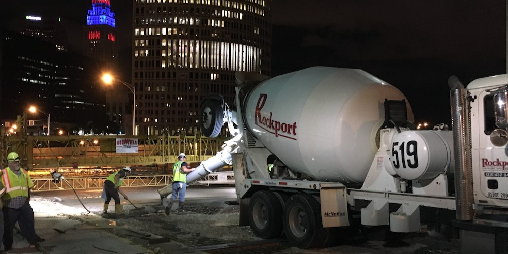 Construction workers pouring concrete at night in front of several street lights and buildings.