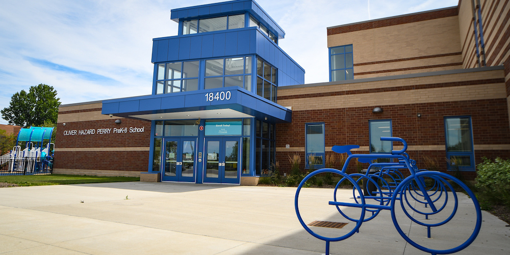 Outside of elementary school made out of mostly dark and light brown brick with blue metal on and above the doors. On the right of the image, bike racks in the shape of blue bikes.