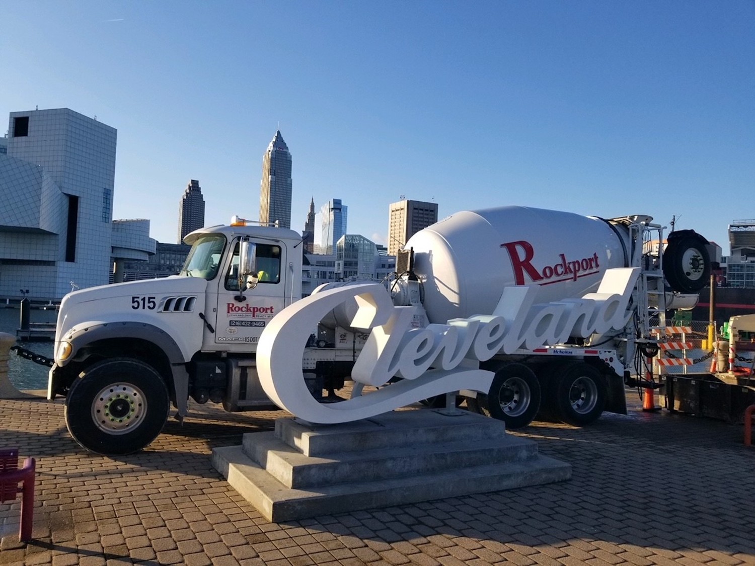 Concrete truck is parked behind white "Cleveland" logo mounted on concrete. Both are in front of city skyline.