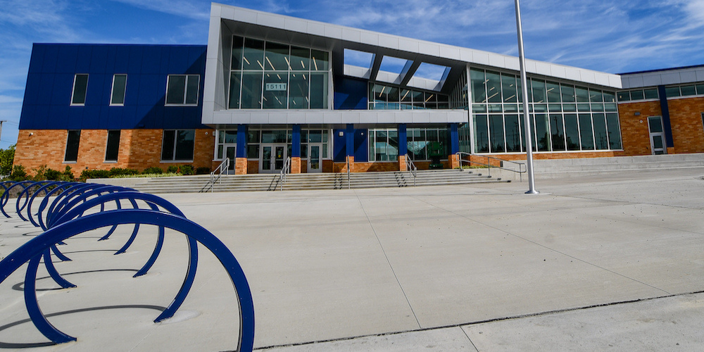 Wide shot of a building entrance with blue bike racks on the left side. The building is dark blue, with brick and metal accents.