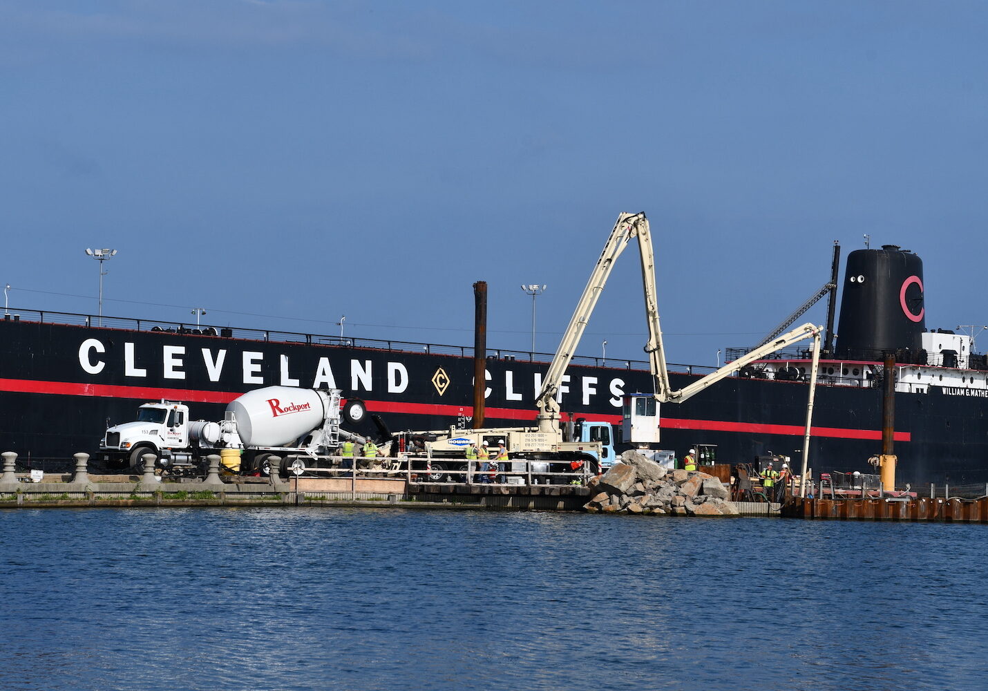 Concrete truck sit in front of black ship with red stripe that reads "Cleveland Cliffs" on the water.
